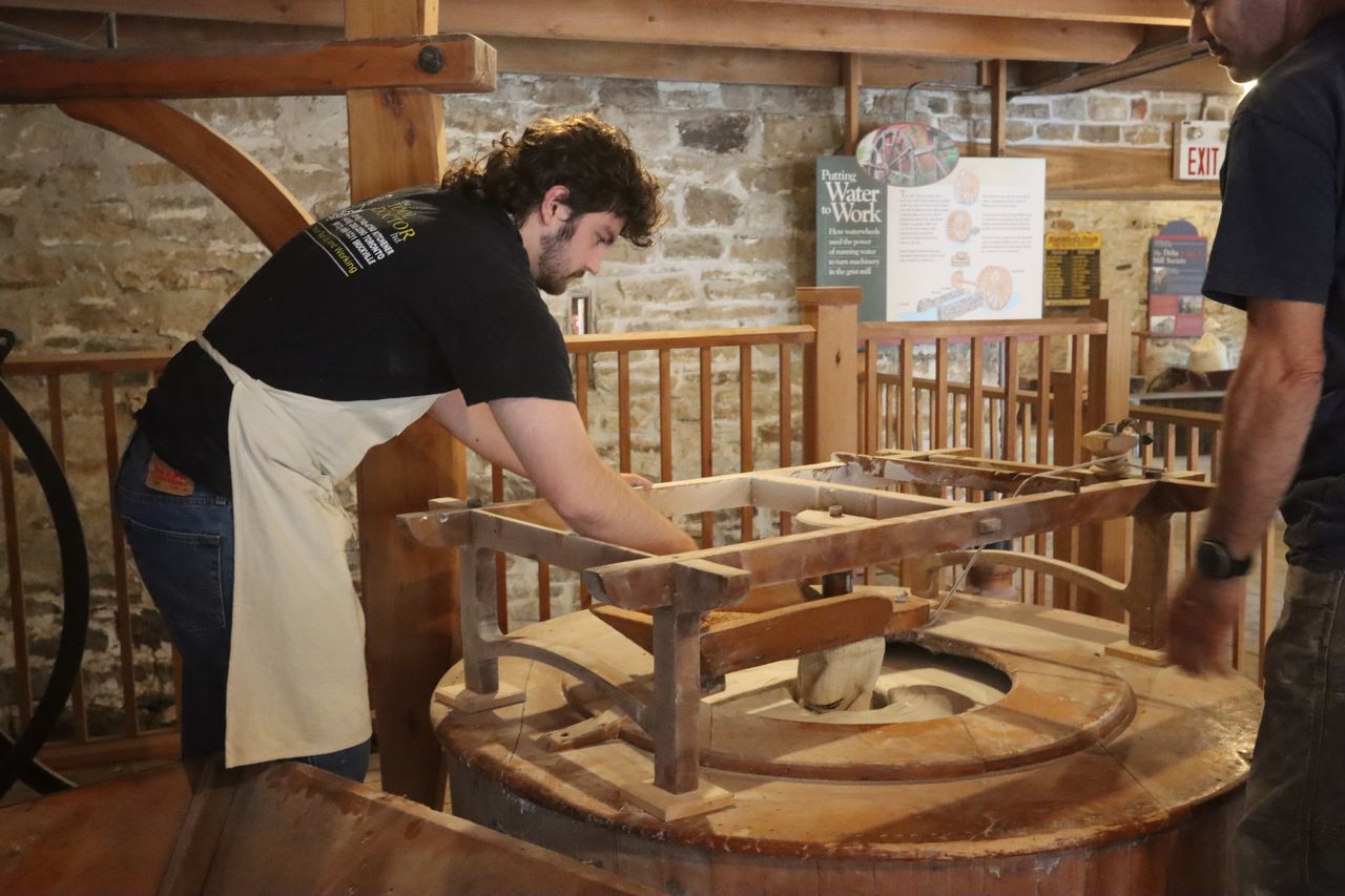 Mill interpreter at the hopper, watching grain fall between the stones